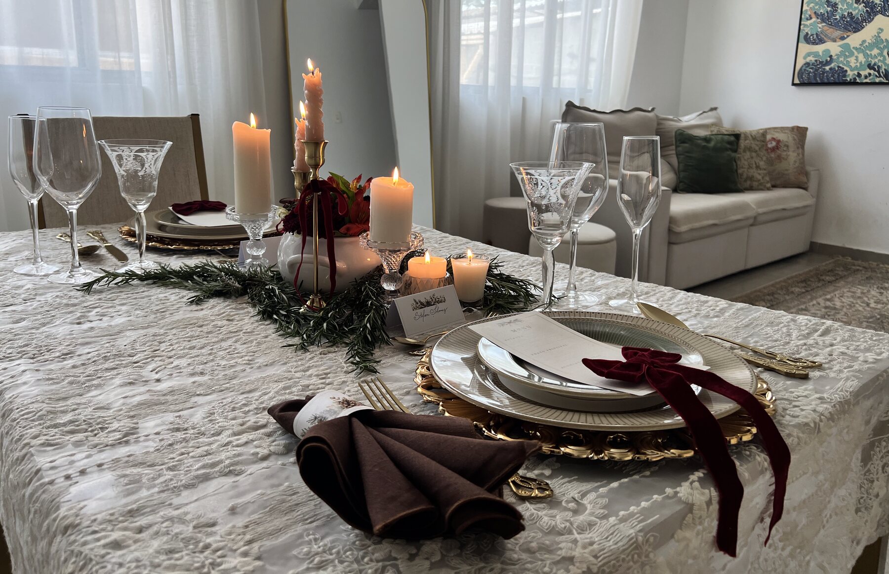 Mesa de Natal posta: toalha rendada, sousplat dourado, guardanapo com laço de veludo e velas acesas ao longo de uma guirlanda de alecrim